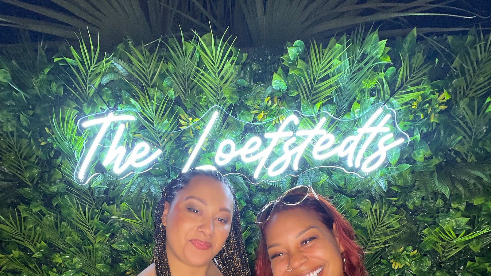 Two guests smiling in front of a custom neon name sign and greenery wall at an Auburn event